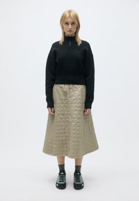 Melnas džemperis ar augstu apkakli un logotipa detaļu, apvienots ar kviltētu bēšas krāsas svārku. Modelis valkā melnas un pelēkas kurpes ar teksturētām zolēm.