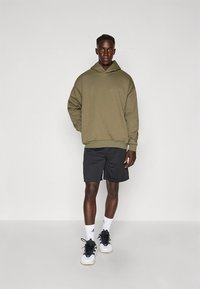 Sudadera de gran tamaño de color verde oliva, pantalones cortos negros, calcetines blancos y zapatillas negras. Tela suave con un ajuste relajado y sin detalles prominentes.