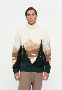 Man wearing a fleece pullover with mountain and pine tree landscape design, standing against a plain white background.