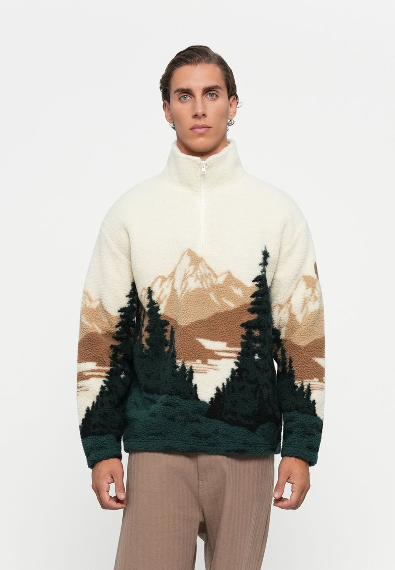 Man wearing a fleece pullover with mountain and pine tree landscape design, standing against a plain white background.