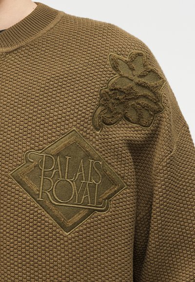Καφέ πλεκτό πουλόβερ με υφή, διακοσμημένο με κεντημένο λουλουδένιο έμβλημα στον ώμο και κείμενο «PALAIS ROYAL» στο στήθος.