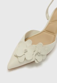 Chaussure slingback blanche en cuir avec un bout pointu, ornée d'accents floraux superposés et d'une texture lisse et mate sur la tige.
