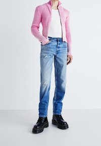 Přiléhavá růžová žebrovaná bunda s vysokým límcem, nošená přes bílý top na ramínka. Modré roztrhané džíny s rovnými nohavicemi a černé robustní boty dokončují vzhled.