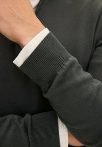 Mörkgrön stickad tröja med ribbade manschetter. Den inre manschetten är ljusbeige, vilket visar kontrast i både textur och färg. Slät tygfinish.