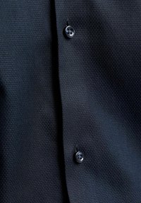 Gros plan d’un tissu noir texturé avec deux boutons noirs attachés, montrant les boutonnières et de subtiles réflexions de lumière sur le matériau.