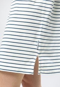 Λευκή φούστα με λεπτές μπλε οριζόντιες ρίγες, μικρή πλαϊνή σχισμή και υφασμάτινη ετικέτα κοντά στο τελείωμα στο πόδι ενός ατόμου.