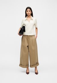 Κρεμ κοντομάνικο πόλο με φερμουάρ, συνδυασμένο με καφέ παντελόνια φαρδιάς γραμμής. Το μοντέλο κρατάει μια μαύρη τσάντα φάκελο και φοράει κόκκινες μυτερές γόβες.