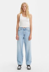 Nainen, jolla on pitkät vaaleat hiukset, pukeutuneena valkoiseen hihattomaan paitaan, vaaleansinisiin leveälahkeisiin farkkuihin ja mustavalkoisiin lenkkareihin, seisoo valkoista taustaa vasten.