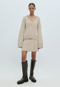 Pull en tricot beige avec un décolleté en V et des manches surdimensionnées, associé à une jupe assortie ; porté avec des bottes hautes marron foncé.