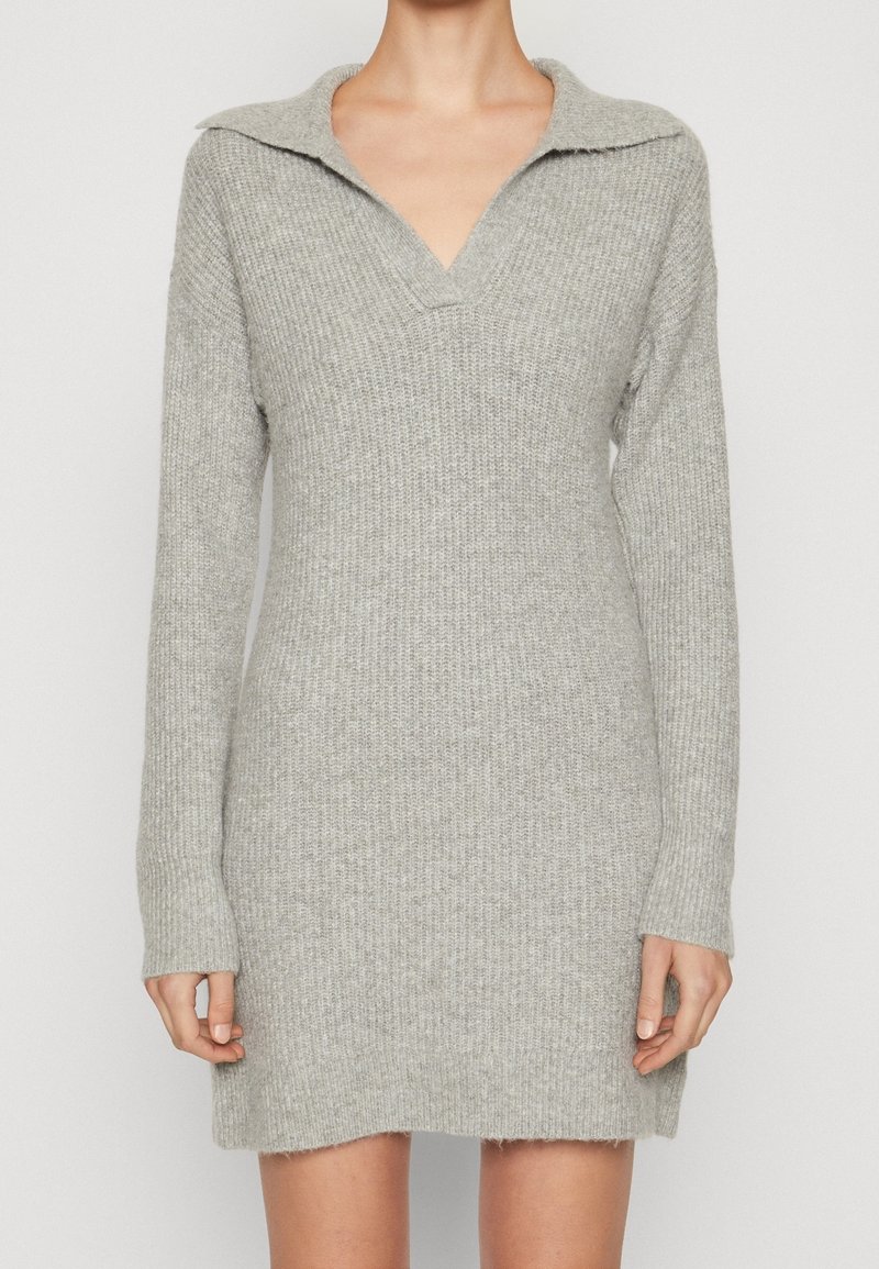 Robe pull en tricot côtelé gris clair avec manches longues et large col en V, portée par une personne debout devant un fond uni.