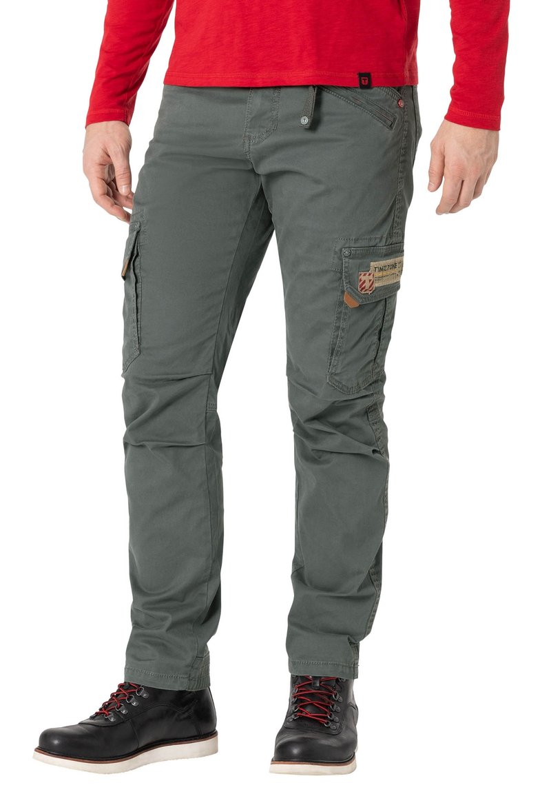 Cargo-Hosen aus olivgrünem Baumwollstoff mit mehreren Taschen, verstärkten Nähten und einem Patch-Detail am Oberschenkel. Kombiniert mit schwarzen Schnürstiefeln.