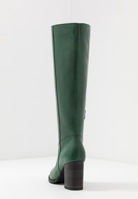 Botte haute jusqu'au genou en cuir vert avec une finition texturée, une forme effilée et un talon en bois solide. Présente des détails de couture et une petite étiquette de logo.