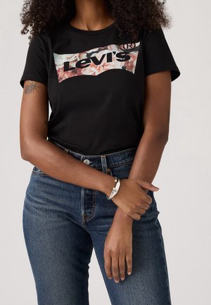 Femme portant un t-shirt noir Levi's avec un logo floral, un jean bleu taille haute, un bracelet argenté et un tatouage sur son bras gauche.