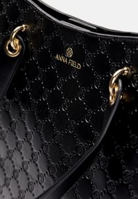 Sac à main en cuir noir avec motifs circulaires embossés, œillets dorés et logo "ANNA FIELD" en lettres dorées.