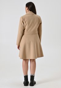 Cappotto di lana beige con una vestibilità sartoriale, maniche lunghe e orlo svasato. La vista posteriore mostra una vita con cintura e un design minimalista. Stivali neri alla caviglia visibili.