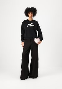 Felpa nera con logo bianco "Hugo", abbinata a pantaloni neri a gamba larga con tasche laterali, che tiene in mano una borsa piccola rosa.