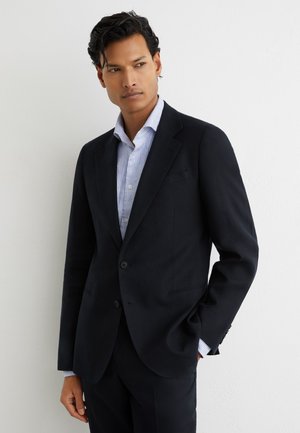 Fekete öltönyzakót és világoskék csíkos inget viselő férfi, egyik kezét a zsebében tartva áll egy sima fehér fal előtt.
