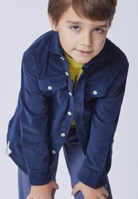 Camicia blu navy con bottoni e due tasche sul petto, realizzata in tessuto morbido. Indossata sopra una t-shirt gialla, abbinata a pantaloni grigi.