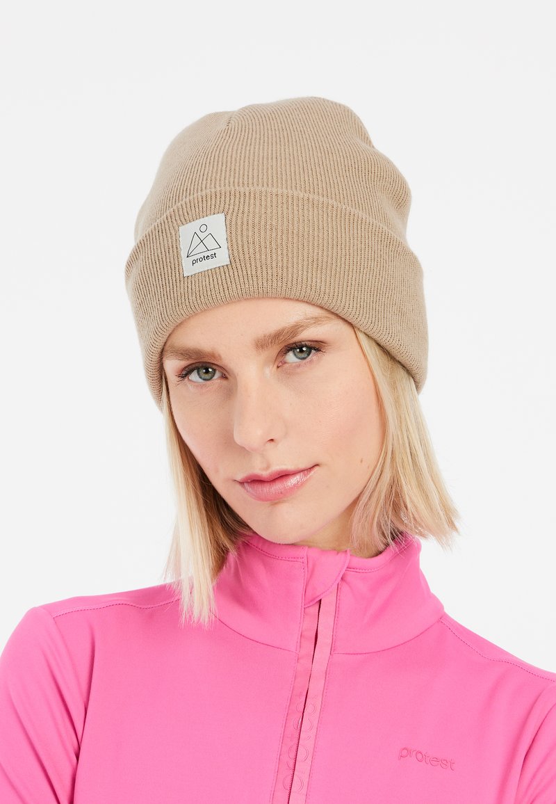 Berretto in maglia color nocciola con texture a coste, dotato di una patch quadrata con logo sul davanti. Il modello indossa un top zip luminoso rosa, con un design aderente.