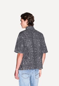 Camicia di jeans nera a maniche corte con motivi floreali e geometrici bianchi, dotata di colletto classico e vestibilità rilassata, indossata con jeans azzurri chiari.