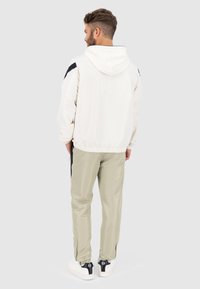 Leichte, crèmefarbene Kapuzenjacke mit schwarzen Akzenten, elastischen B ündchen und lässiger Passform, kombiniert mit beigen Hosen und weißen Sneakers.