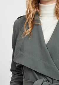Cappotto grigio avvolgente con colletto drappeggiato, ampi rever e cintura. Indossato sopra un dolcevita bianco, realizzato in tessuto morbido e texturizzato.