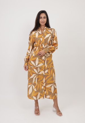 Robe maxi en tissu à motifs présentant des designs de feuilles marron, jaune et blanc. Manches longues et fermeture à boutons sur le devant avec une taille cintrée. Talons transparents.