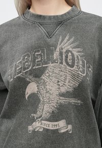 Felpa grigia con una stampa grafica di un'aquila e la parola "REBELLIOUS" in uno stile sbiadito e usurato. Presenta un colletto a V.