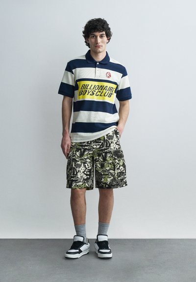 Νέος άνδρας στέκεται φορώντας μπλε-λευκή ριγέ πόλο με το κείμενο "Billionaire Boys Club", κοντά καμουφλάζ παντελόνια cargo, γκρι κάλτσες και μαύρα λευκά αθλητικά παπούτσια.