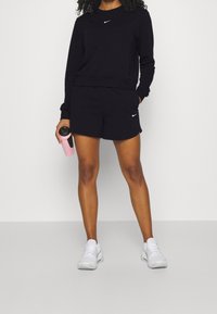 Musta Nike-huppari ja shortsit, joissa on logo rinnassa ja reidessä, yhdistettynä valkoisiin lenkkarikenkiin ja vaaleanpunaiseen vesipulloon.