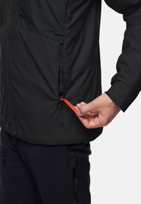 Schwarze Jacke mit einer Reißverschlusstasche; kontrastierendes dunkles Stoffpanel auf der Brust. Hand, die einen orangen Zugriegel an der Taille anpasst.