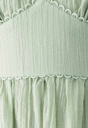 Light green fabric with a textured finish, featuring a ruffled hem and circular detailing along the seam. Subtle sheen and soft drape.