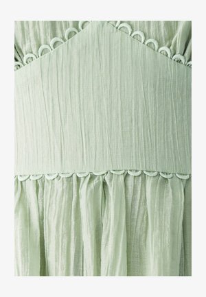 Light green fabric with a textured finish, featuring a ruffled hem and circular detailing along the seam. Subtle sheen and soft drape.
