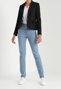 Veste noire avec revers, bouton devant et deux poches, associée à un jean skinny bleu clair et des baskets blanches. Texture plate et lisse sur toute la surface.