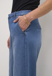 Ljust blå denimbyxor med vida ben och framfickor. Har kontrasterande sömmar och en avslappnad design. Bärs med ett guldarmband.