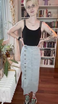 Persoon met kort blond haar en tatoeages draagt een zwart tanktop, een lichte denimrok met knopen en kleurrijke sneakers in een kamer met boekenplanken.