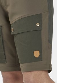 Lähedalt vaadatuna oliivroheliste cargo-lühikesed püksid taskul, kus on nööbid ja väike nahkline vertikaalne logoäpud mudeli reie peal.