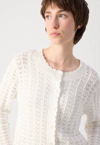 Hvit strikket cardigan med et teksturert mønster, ribbestrikket hals og myke knapper foran. Lett og pustende design.
