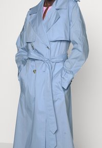 Jasnoniebieski płaszcz trench z podwójnym zapięciem, dużym kołnierzem, paskiem w talii i bocznymi kieszeniami. Gładka faktura materiału z minimalnymi detalami.
