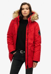 Chaqueta parka roja con capucha forrada de piel, cierre de cremallera y botones a presión, puños acanalados y un parche en la manga izquierda. Lleva encima de un suéter negro.