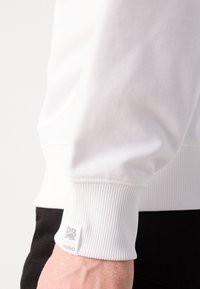 Hvit bomullssweatshirt med ribbede mansjetter og en subtil logoetikett. Strukturen er glatt og passformen ser avslappet ut.