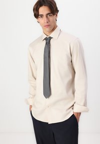 Chemise longue beige clair à manches longues avec un col boutonné, associée à une cravate texturée gris foncé, portée avec un pantalon marine. Tissu doux, coupe ajustée.