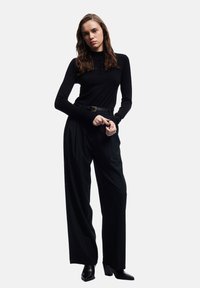 Frau mit langen braunen Haaren, die ein schwarzes Langarmoberteil mit Knöpfen, weite schwarze Hosen, einen schwarzen Gürtel und schwarze hohe Schuhe trägt und steht.