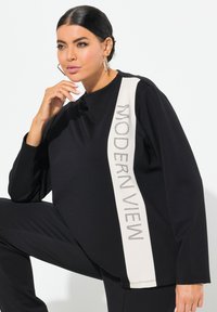 Maglietta nera con maniche lunghe, caratterizzata da una striscia beige sul lato con la scritta "MODERN VIEW" in strass. Tessuto morbido con una vestibilità rilassata.