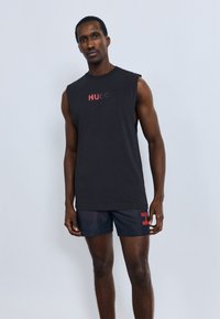 Črna majica brez rokavov z napisom "HUGO" v rdeči barvi, kombinirana s temnimi kratkimi hlačami z rdečim grafičnim poudarkom na strani. Gladek bombažni material.