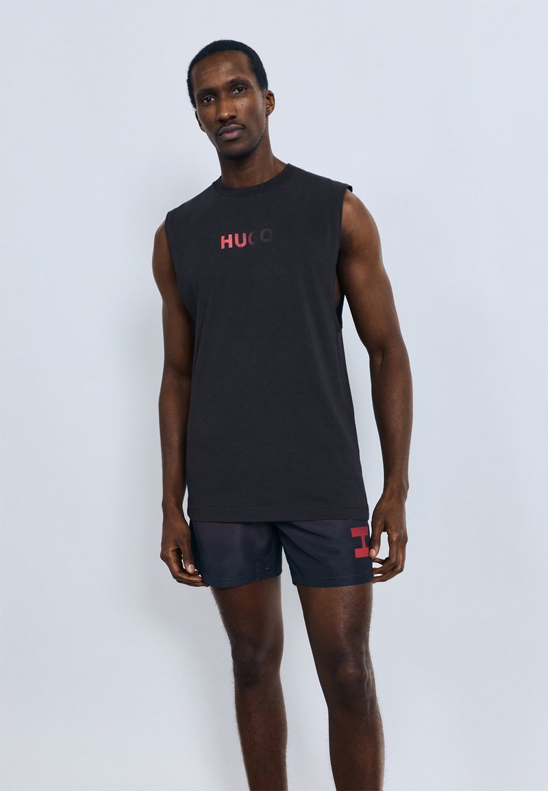 Črna majica brez rokavov z napisom "HUGO" v rdeči barvi, kombinirana s temnimi kratkimi hlačami z rdečim grafičnim poudarkom na strani. Gladek bombažni material.