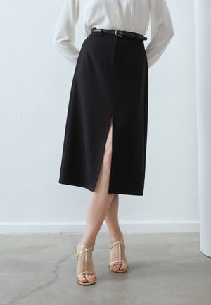 Personne portant une jupe midi noire avec une fente avant, un chemisier blanc à manches longues, une ceinture noire et des sandales à talons hauts à lanières dorées.