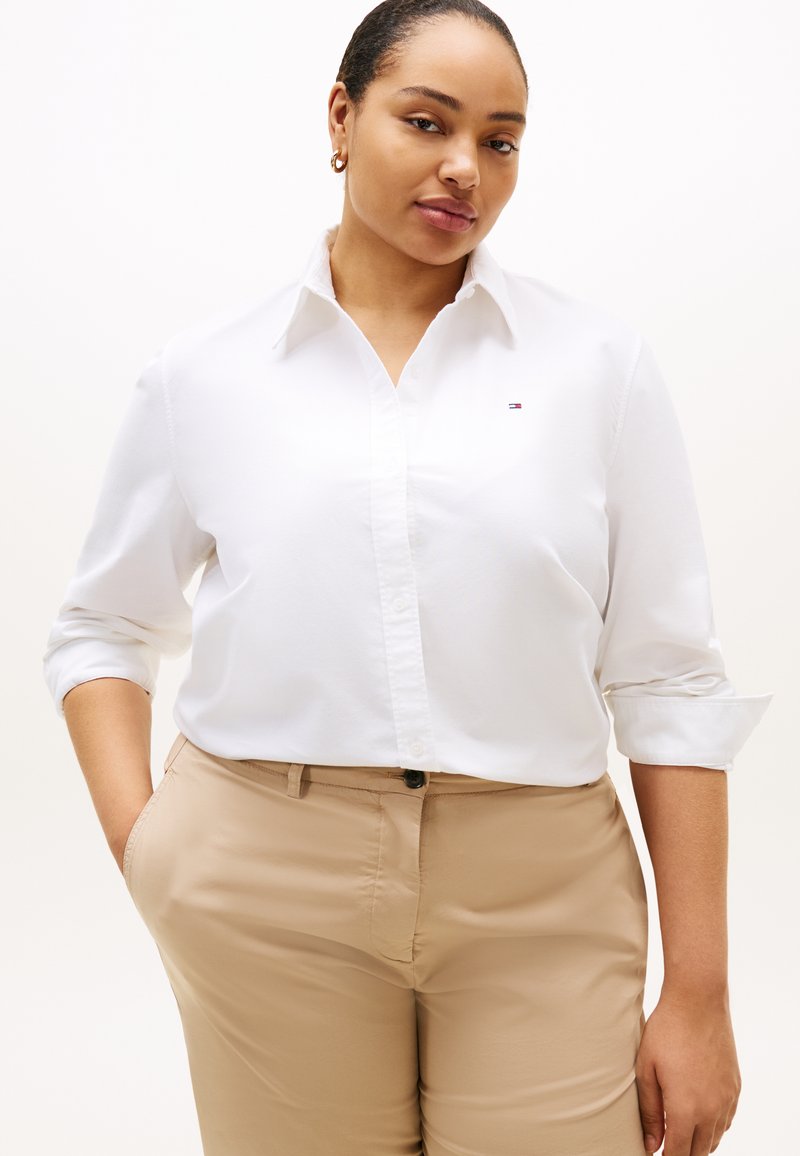 Λευκό πουκάμισο με κουμπιά και ρεβέρ στα μανίκια, με μικρό λογότυπο στο στήθος· συνδυασμένο με ανοιχτό μπεζ παντελόνι. Υφασμάτινο με ομαλή υφή, εφαρμοστό σχέδιο.