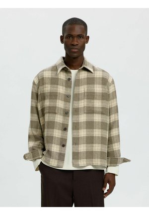 Homme portant une veste chemise à carreaux beige et marron par-dessus une chemise blanche à manches longues et un pantalon foncé, debout devant un fond uni.