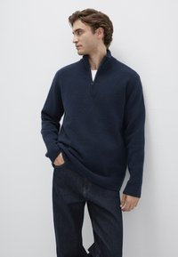 Navy fleece sweater with a quarter-zip collar, long sleeves, and a relaxed fit, paired with dark jeans. Soft texture and minimal design.